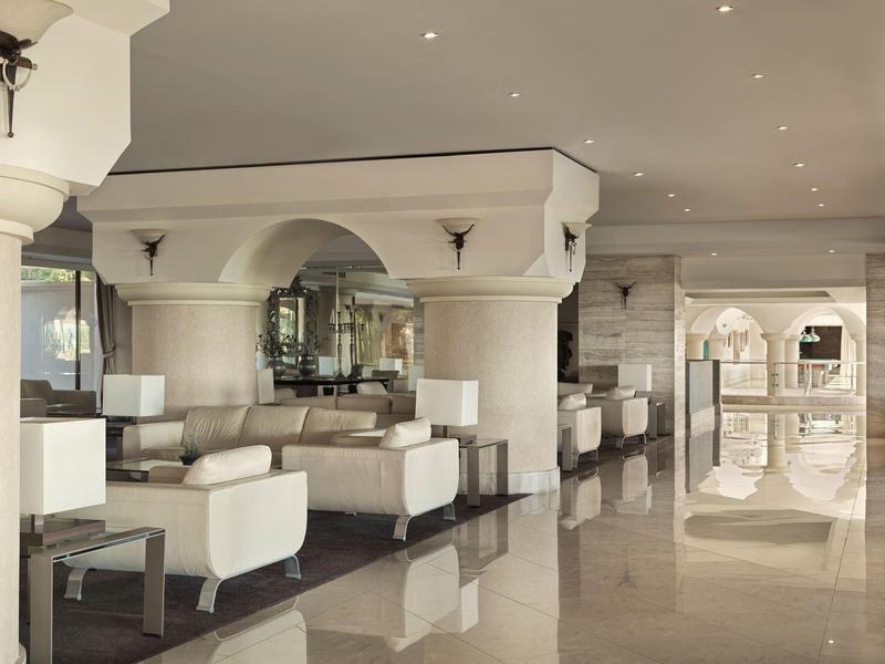 Area lounge d'hotel elegante e luminosa con poltrone bianche e pavimento riflettente.