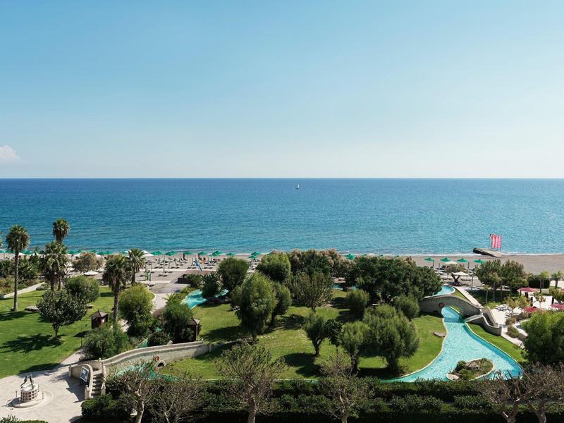 Vista sul mare con prato verde, alberi, piscina e spiaggia soleggiata.