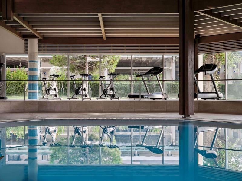 Spazio interno con piscina e attrezzature fitness, circondato da grandi finestre con vista sul verde.