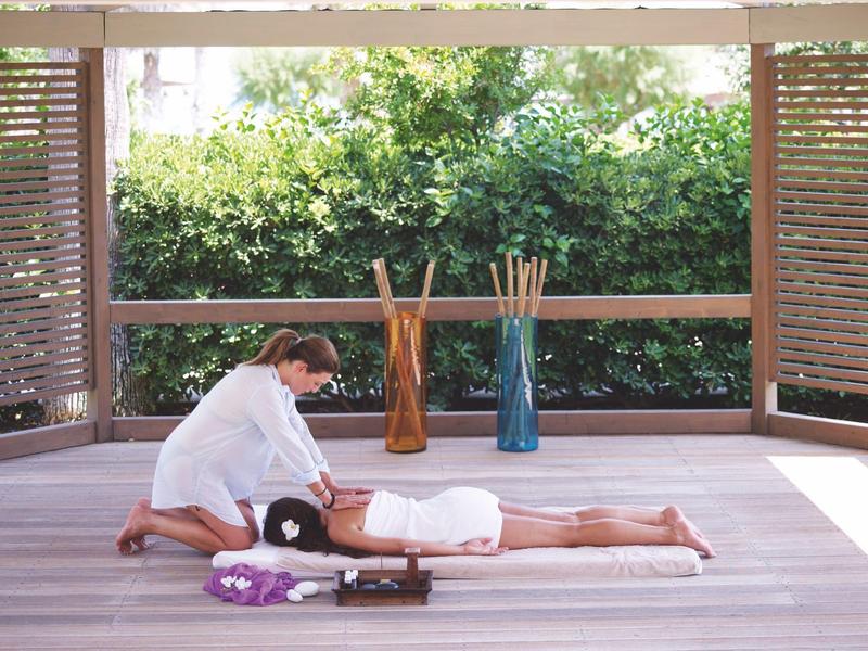 Una donna riceve un massaggio su una terrazza in legno con piante verdi sullo sfondo.