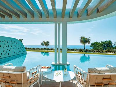 Area piscina rilassante con sedie comode, palme e vista sull'oceano sotto una pergola.