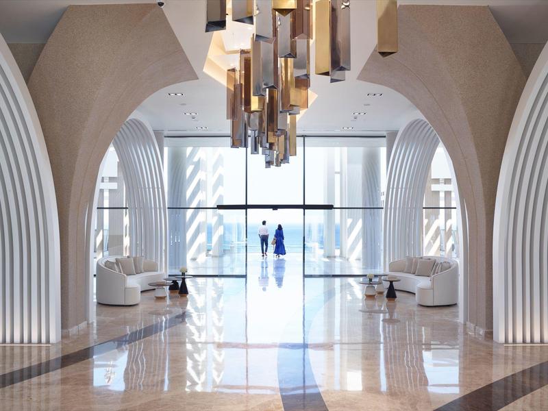 Lobby di hotel moderno con grandi archi, lampadario e pavimento lucido che riflette la luce.