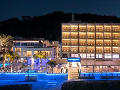 Oświetlony hotel z basenem i palmami nocą, otoczony spokojnym krajobrazem.