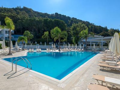 Piscina moderna de hotel con tumbonas y palmeras frente a colina boscosa bajo cielo azul.