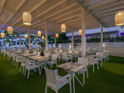 Oświetlony zewnętrzny teren restauracji z białymi stołami i krzesłami na zielonej podłodze.