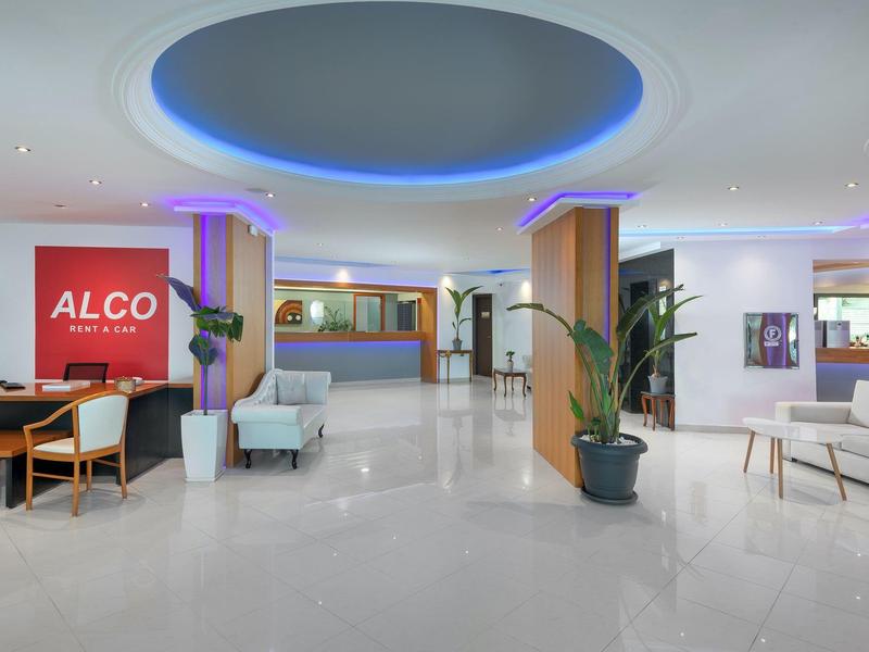 Lobby de hotel luminosa con muebles modernos, recepción y grandes plantas en tonos blancos y morados.