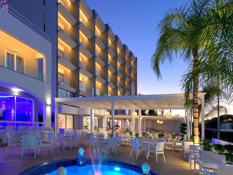 Vista nocturna de un hotel iluminado con piscina, mobiliario blanco y palmeras.