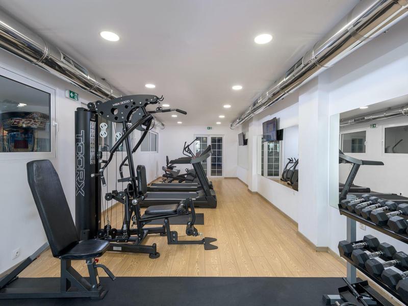 Gimnasio moderno con máquinas y pesas, paredes claras, suelo de madera y pared de espejos.