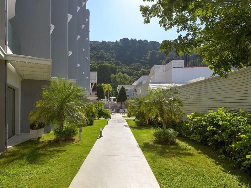 Camino a través de un jardín con palmeras y arbustos junto a un edificio moderno bajo cielo azul.