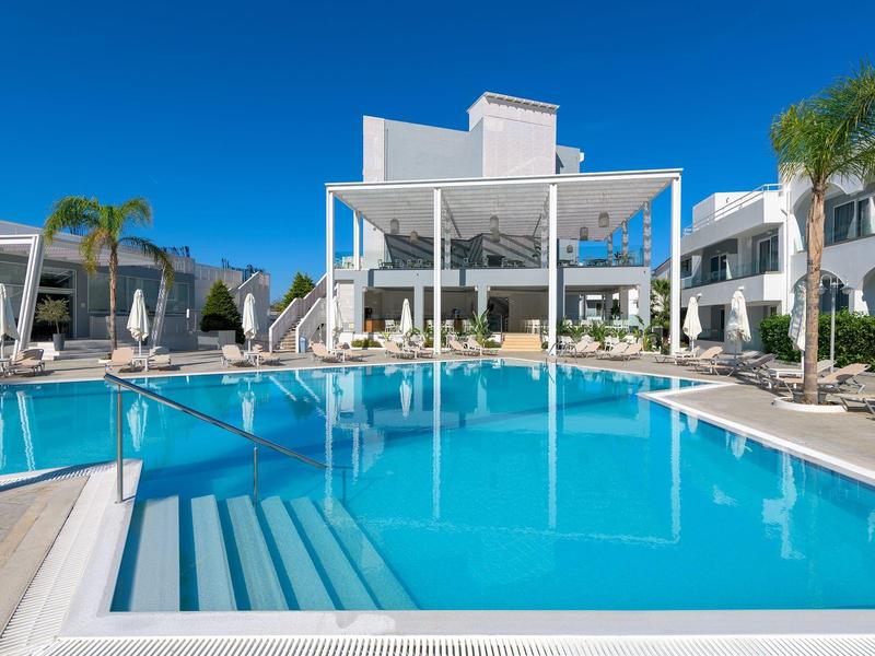 Complejo hotelero blanco moderno con gran piscina clara y tumbonas bajo cielo azul.
