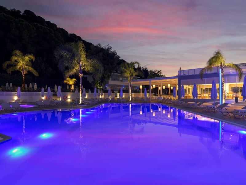 Área de piscina iluminada al atardecer con palmeras y tumbonas en un hotel.