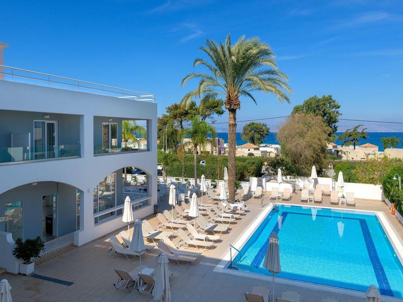 Jardín del hotel con piscina, tumbonas y palmeras, vista al mar azul bajo cielo despejado.