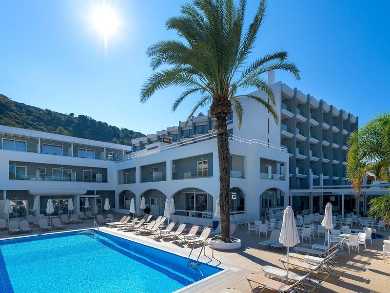 Edificio de hotel con piscina azul, tumbonas y palmeras bajo un cielo despejado.