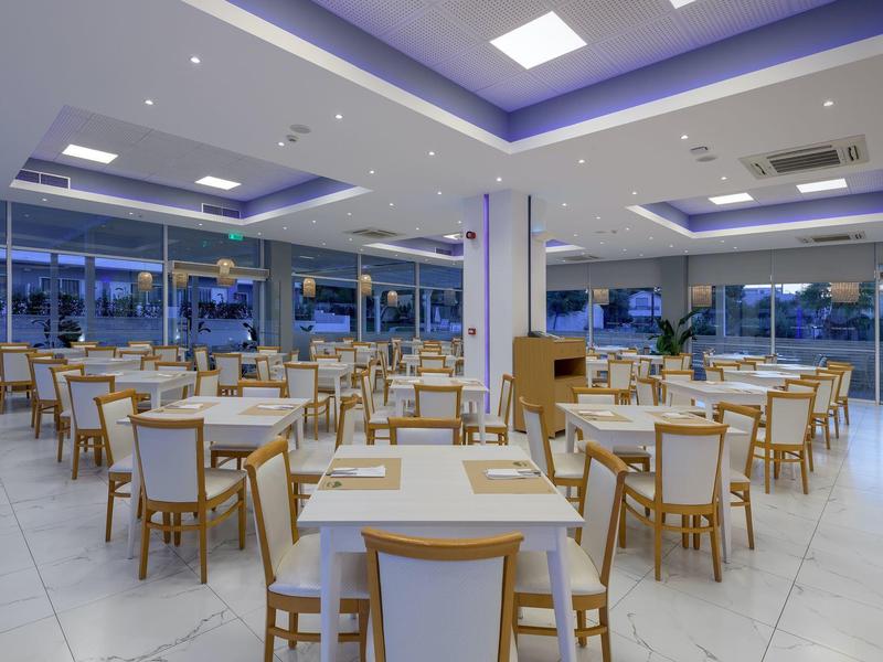 Amplio comedor moderno con mesas y sillas blancas, suelo claro y grandes ventanas.