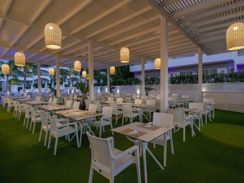 Zona exterior de un restaurante con sillas blancas y linternas colgantes al anochecer.