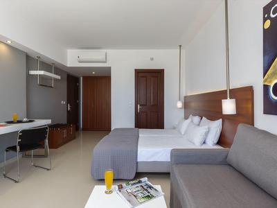 Helles Hotelzimmer mit Doppelbett, grauer Couch, Schreibtisch, Holztüren und moderner Wandkunst.