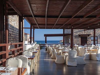 Offenes Restaurant mit weißen Stühlen und Holzdach, Blick auf das Meer im Hintergrund bei klarem Himmel.