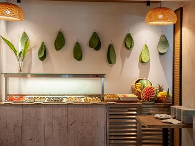 Buffet mit Obst und warmem Essen, Dekoration aus großen grünen Blättern an weißer Wand.