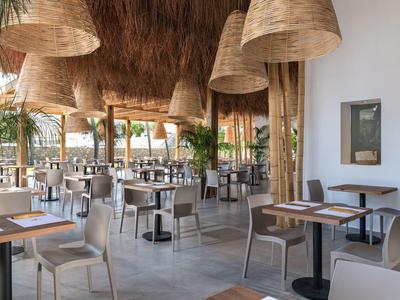 Moderner Außenbereich eines Restaurants mit Strohdach, Holzsäulen, weißen Tischen und grauen Stühlen.