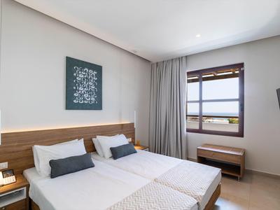 Helles Hotelzimmer mit zwei Einzelbetten, Holzmöbeln, grauem Vorhang und Fenster mit Blick nach draußen.