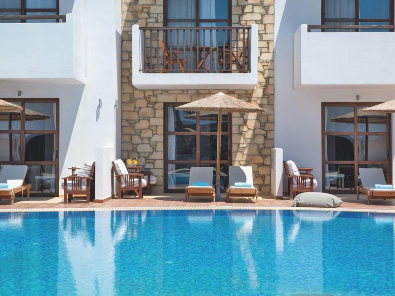 Piscina exterior frente a un hotel con tumbonas y sombrillas en la terraza.