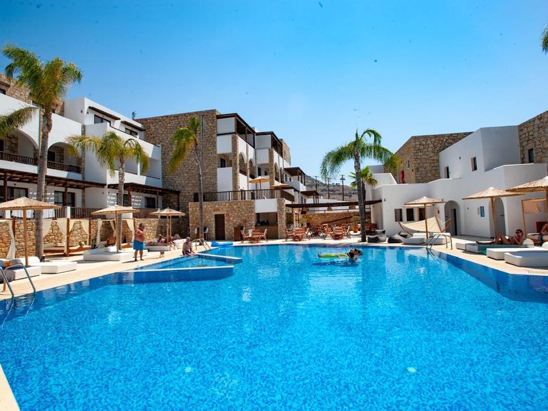 Gran piscina azul de hotel rodeada de tumbonas, palmeras y edificios modernos de varios pisos.