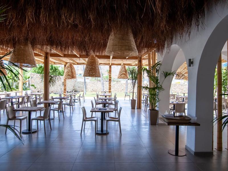 Área de restaurante abierta y luminosa con techo de madera y mesas y sillas blancas en resort tropical.
