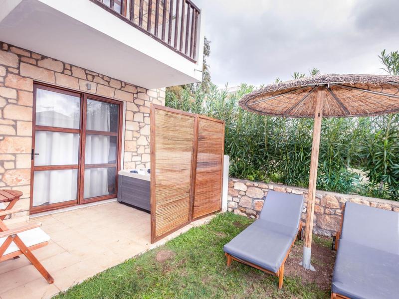 Terraza de hotel con dos tumbonas, sombrilla de paja, mesa y sillas junto a un muro de piedra y césped