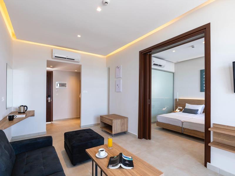 Habitación de hotel moderna con área de sala, sofá, mesa de centro y dormitorio contiguo con cama doble.