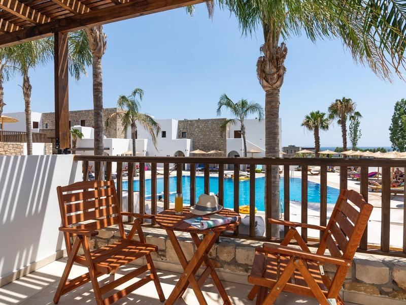 Balcón con mesa y sillas de madera con vista a una piscina y palmeras en un día soleado.