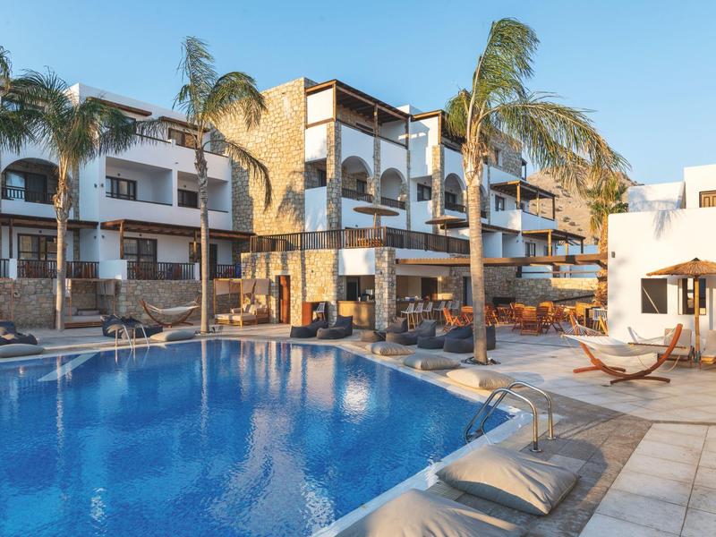 Hotel con piscina exterior, tumbonas y palmeras bajo un cielo azul claro.