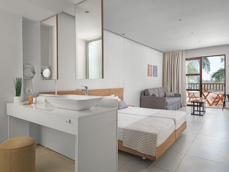 Habitación de hotel moderna con cama, sofá y balcón con vista a palmeras.