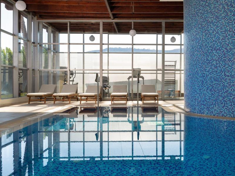 Piscina interior con paredes de mosaicos azules, tumbonas y grandes ventanas con vista a las montañas.