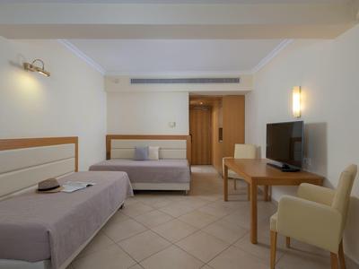 Eenvoudige hotelkamer met twee eenpersoonsbedden, een bureau, tv en beige fauteuils, helder en minimalistisch.