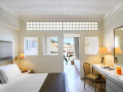 Licht hotelkamer met groot bed, stoelen, lampen en balkon met uitzicht op de kust.