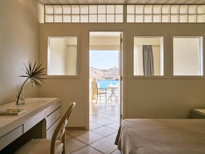 Hotelkamer met open deur naar balkon, zeezicht, bureau, stoel en bed in warm licht.