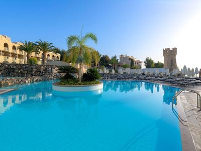 Großer klarer Pool mit Palmeninsel, Liegestühlen und beigen Gebäuden im sonnigen Ferienort.