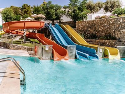 Kleurrijke glijbanen naast een zwembad bij een zonnig resort met stenen muren en groen.