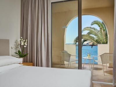 Hotelkamer met balkon met uitzicht op zwembad, palmbomen en oceaan onder een heldere lucht.