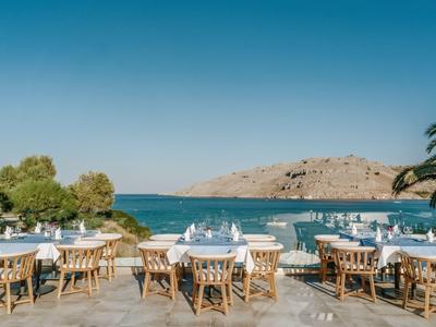 Restaurant vide avec tables en bois au bord de l'eau et vue sur les collines sous un ciel clair.