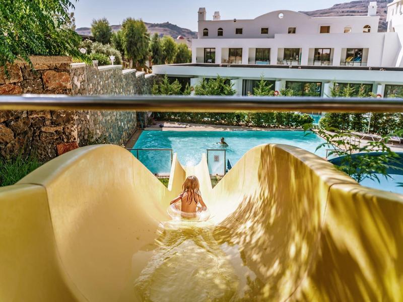 Gele waterglijbaan met uitzicht op een zwembad en witte hotelgebouwen met bergen op de achtergrond.