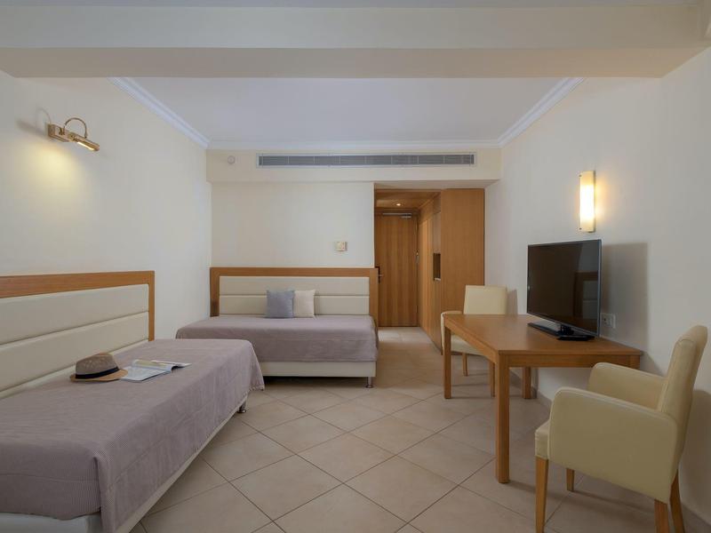 Eenvoudige hotelkamer met twee eenpersoonsbedden, een bureau, tv en beige fauteuils, helder en minimalistisch.