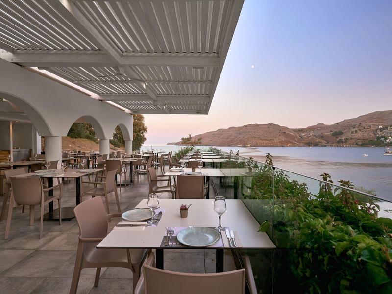 Buiten eetgedeelte met tafels en stoelen met uitzicht op een rustige zee en heuvels bij zonsondergang.