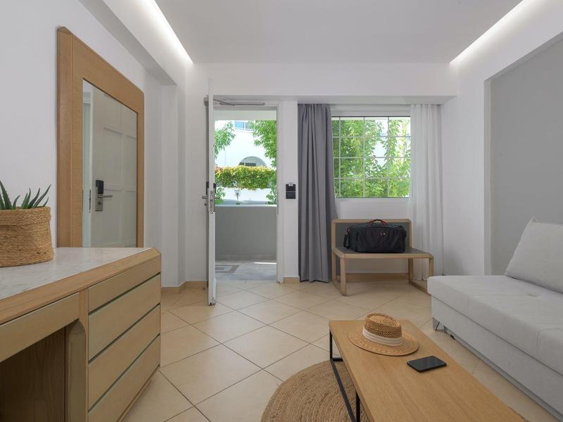 Minimalistische hotelkamer met bank, houten bureau, fauteuil en balkon met uitzicht op groen.