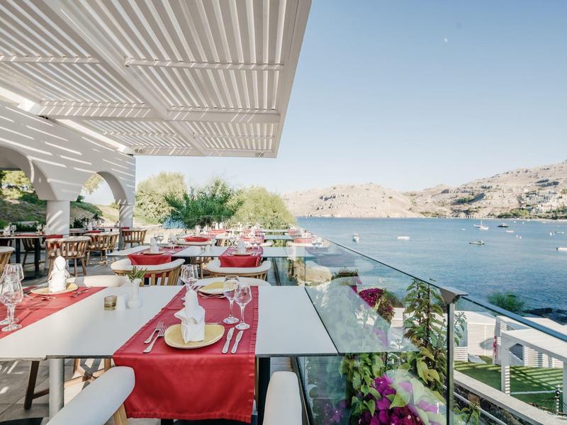 Buitenterras van een restaurant met rode tafelkleden met uitzicht op een kalme blauwe zee en rotsachtig landschap.