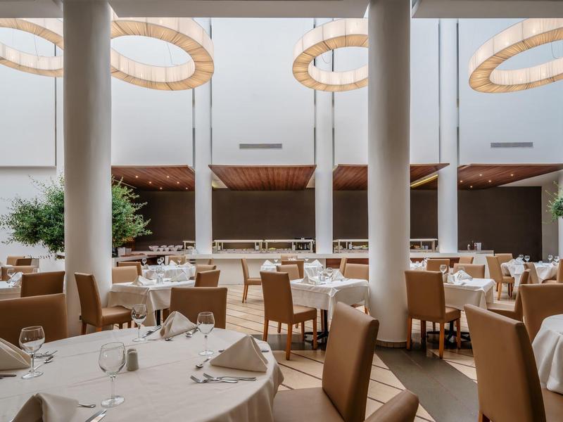 Ruime hotel eetzaal met ronde tafels, beige stoelen, grote zuilen en ronde plafondlampen.