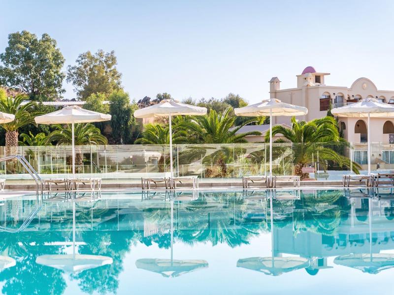 Helderblauw zwembad met witte parasols en een zonnig hotel in mediterrane stijl op de achtergrond.