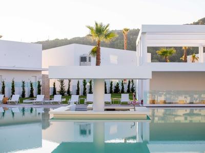 Moderner weißer Gebäudekomplex mit rechteckigem Pool, Palmen und Sitzbereichen bei Tageslicht.