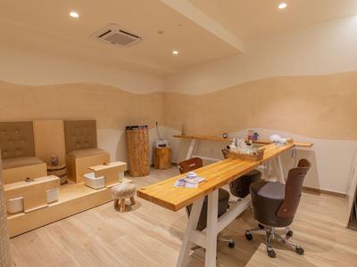 Heller Behandlungsraum mit Massageliege, Holzbank und Rollstuhl, beige Wände und Holzboden.