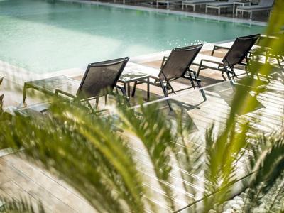 Schwarze Liegestühle am hellen Pool mit ruhigem Wasser, grüne Palmenblätter im Vordergrund.